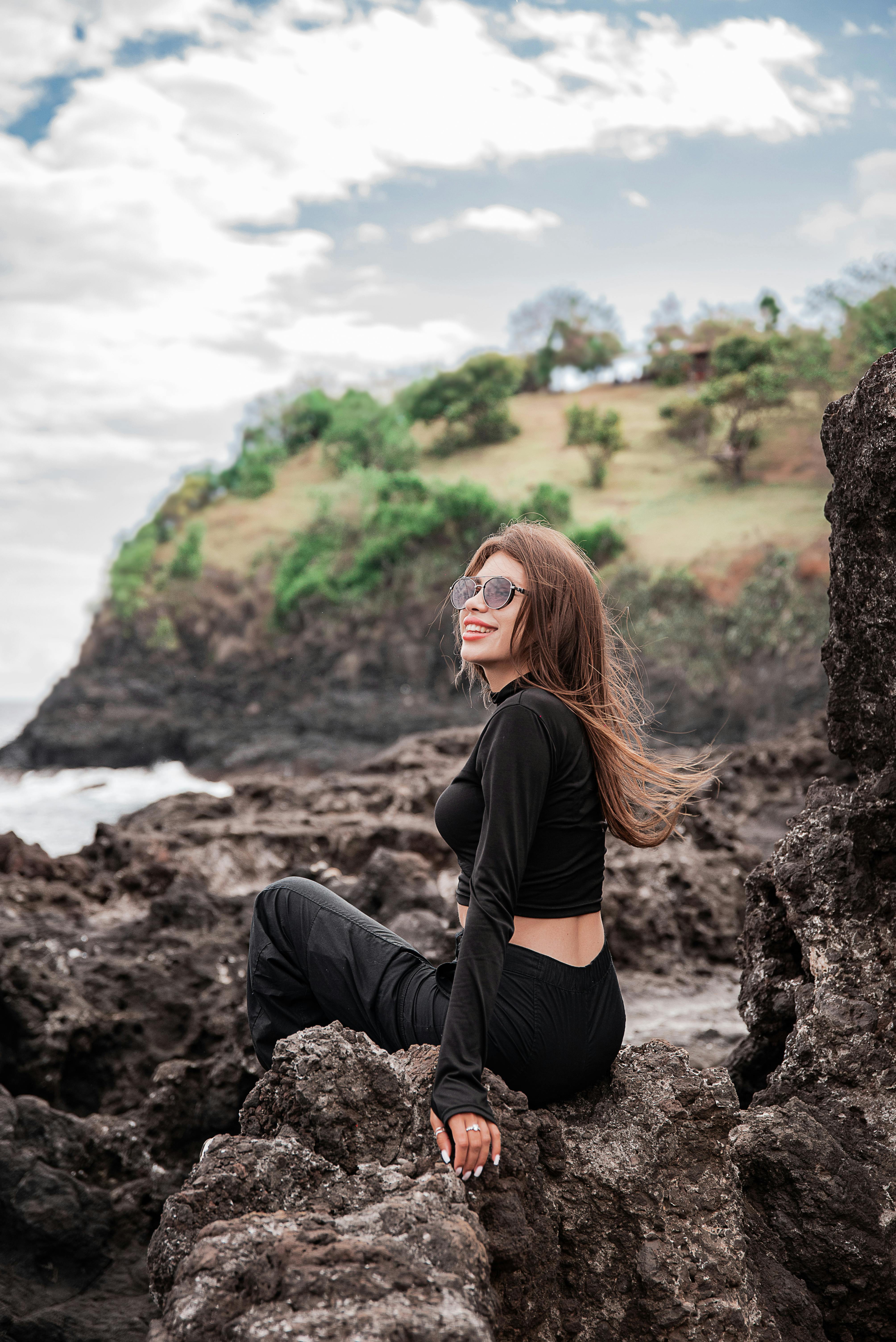 在一个晴朗的日子里，一位年轻女子穿着黑色服装坐在岩石海岸线上，背景是悬崖