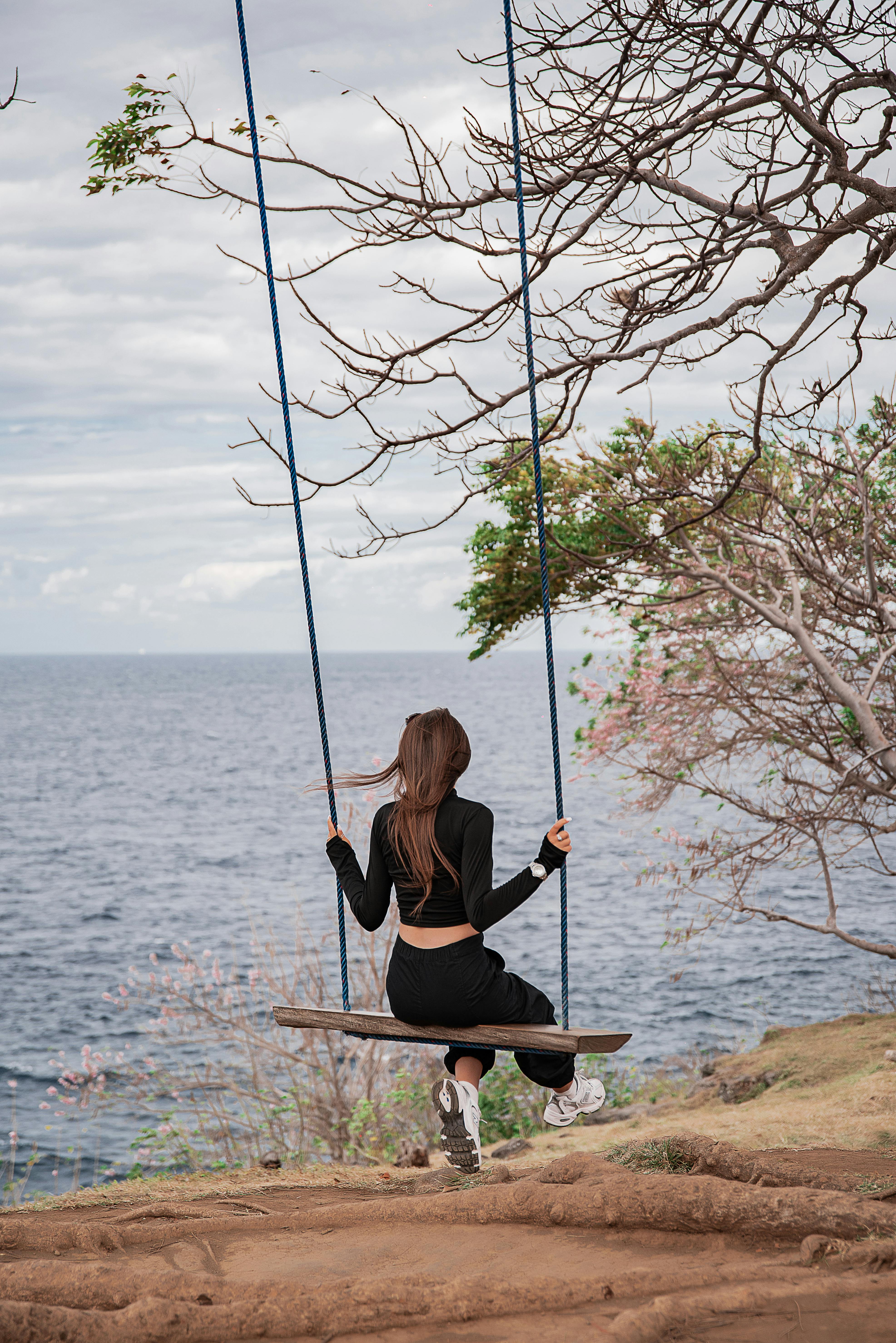 一位穿着休闲装的女士在宁静的海洋背景下荡秋千，周围环绕着海岸树木