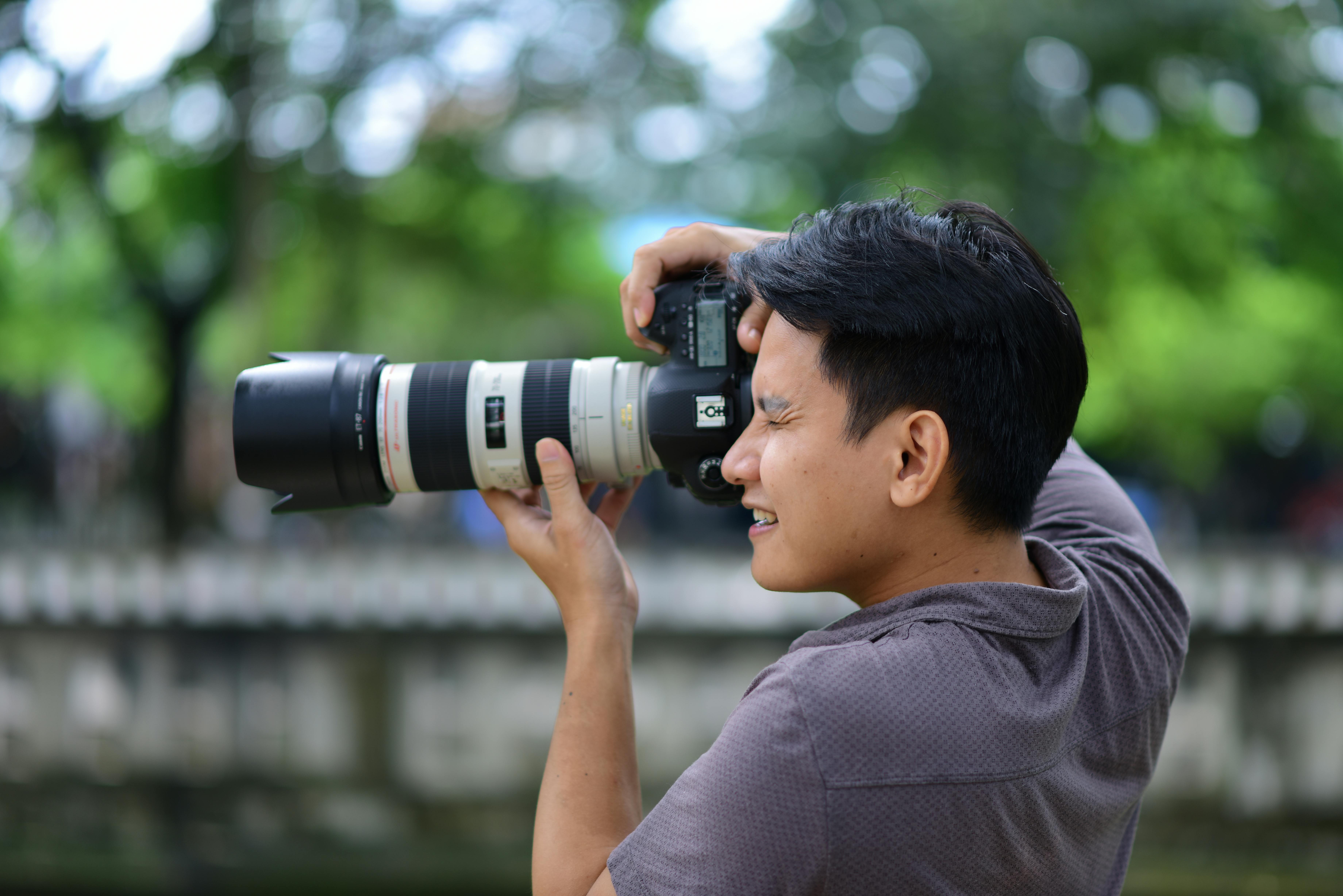 Free A focused photographer using a DSLR camera with a large lens outdoors against a green backdrop. Stock Photo
