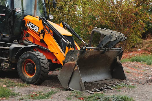 A brightly colored excavator machine parked on a worksite surrounded by autumn trees.