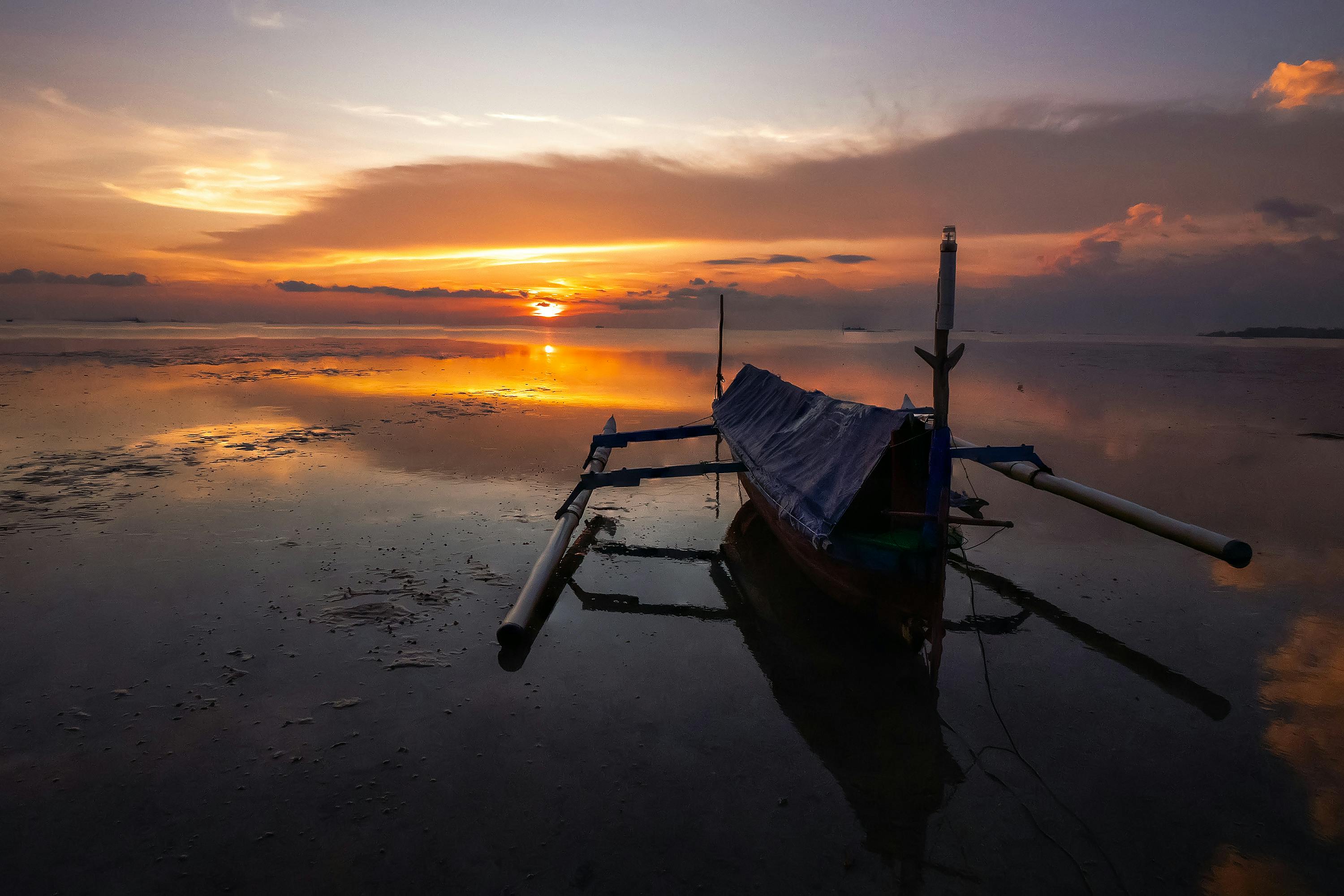 在印度尼西亚邦加勿里洞，一艘传统船只在日落时倒映在平静的水面之上，呈现出宁静而美丽的景色