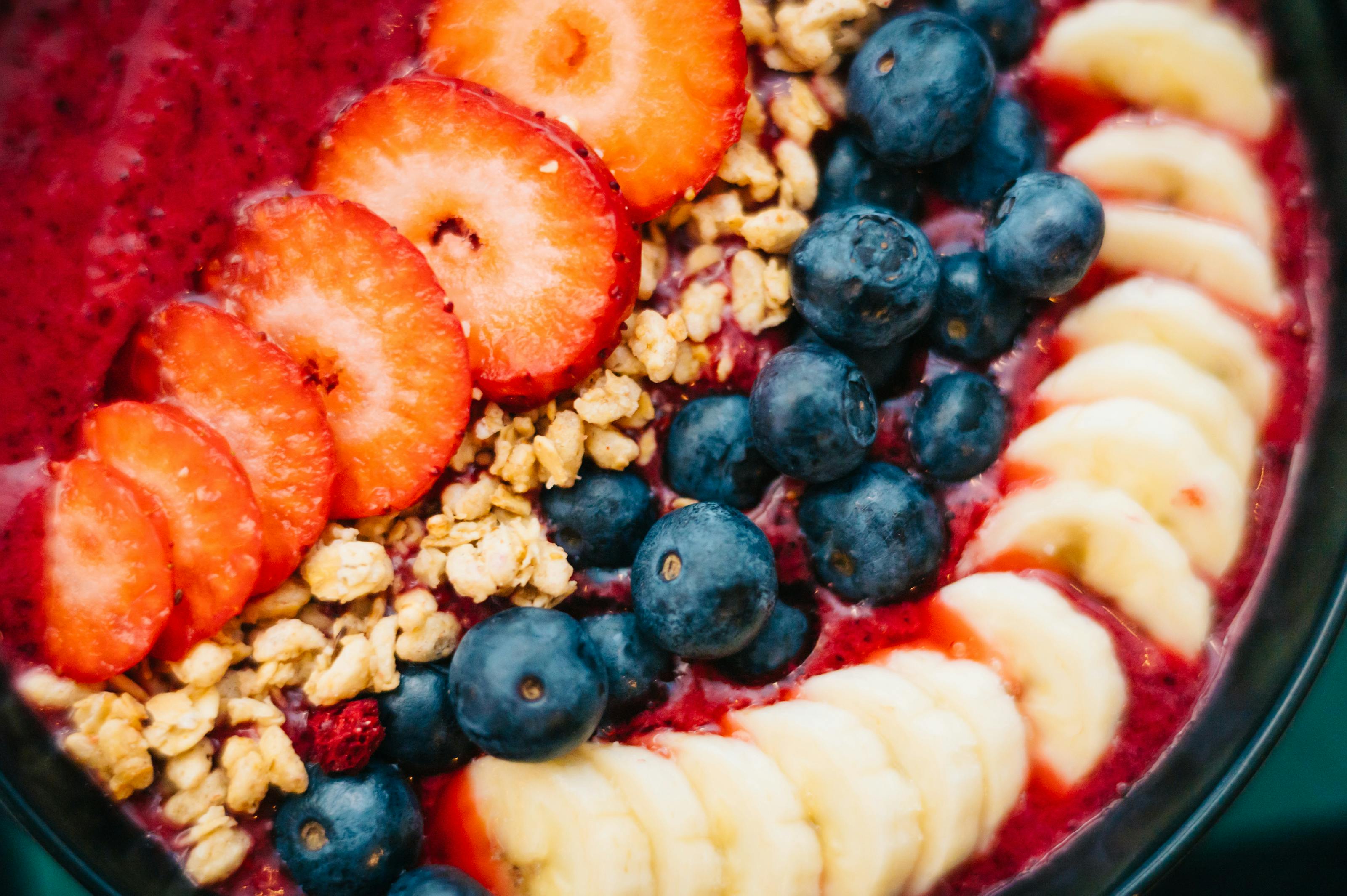 Stack of vegan pancakes with berries and banana, smoothie jar, and fresh fruit bowl on kitchen counter in sunlight.