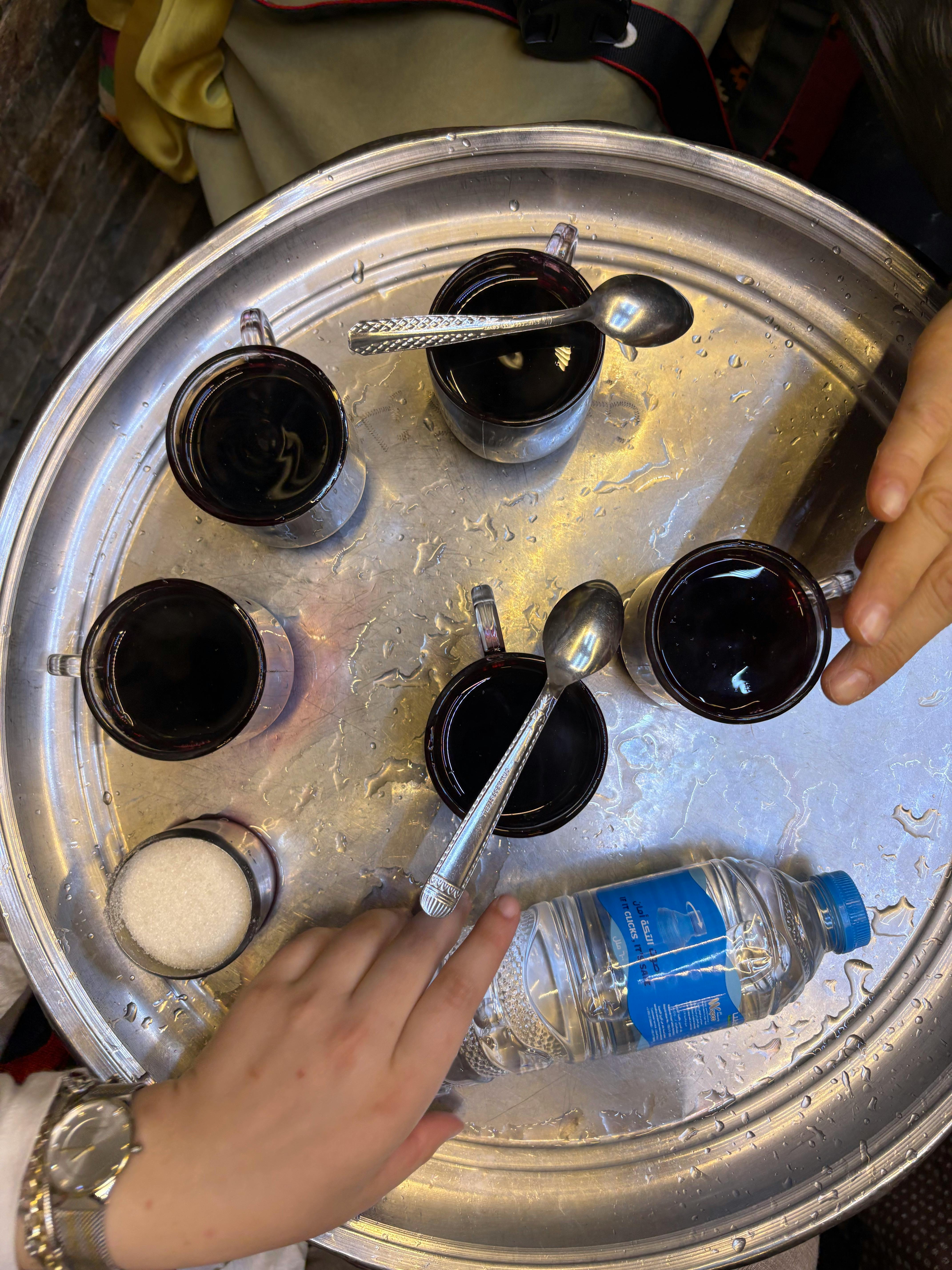 Overhead view of hands serving tea on a metal tray with spoons and water bottle.