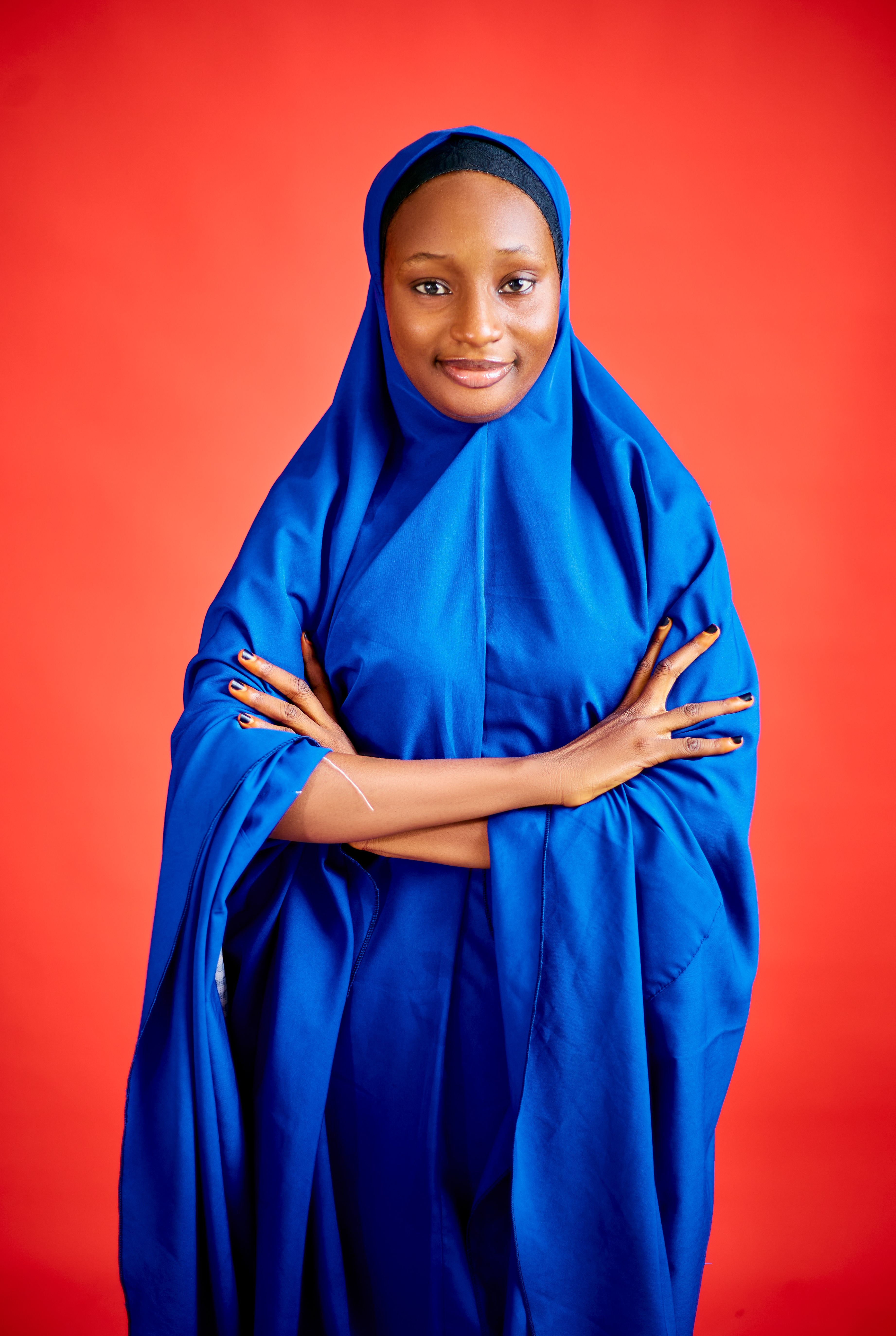 Woman in traditional blue hijab, smiling confidently against a bright red background.