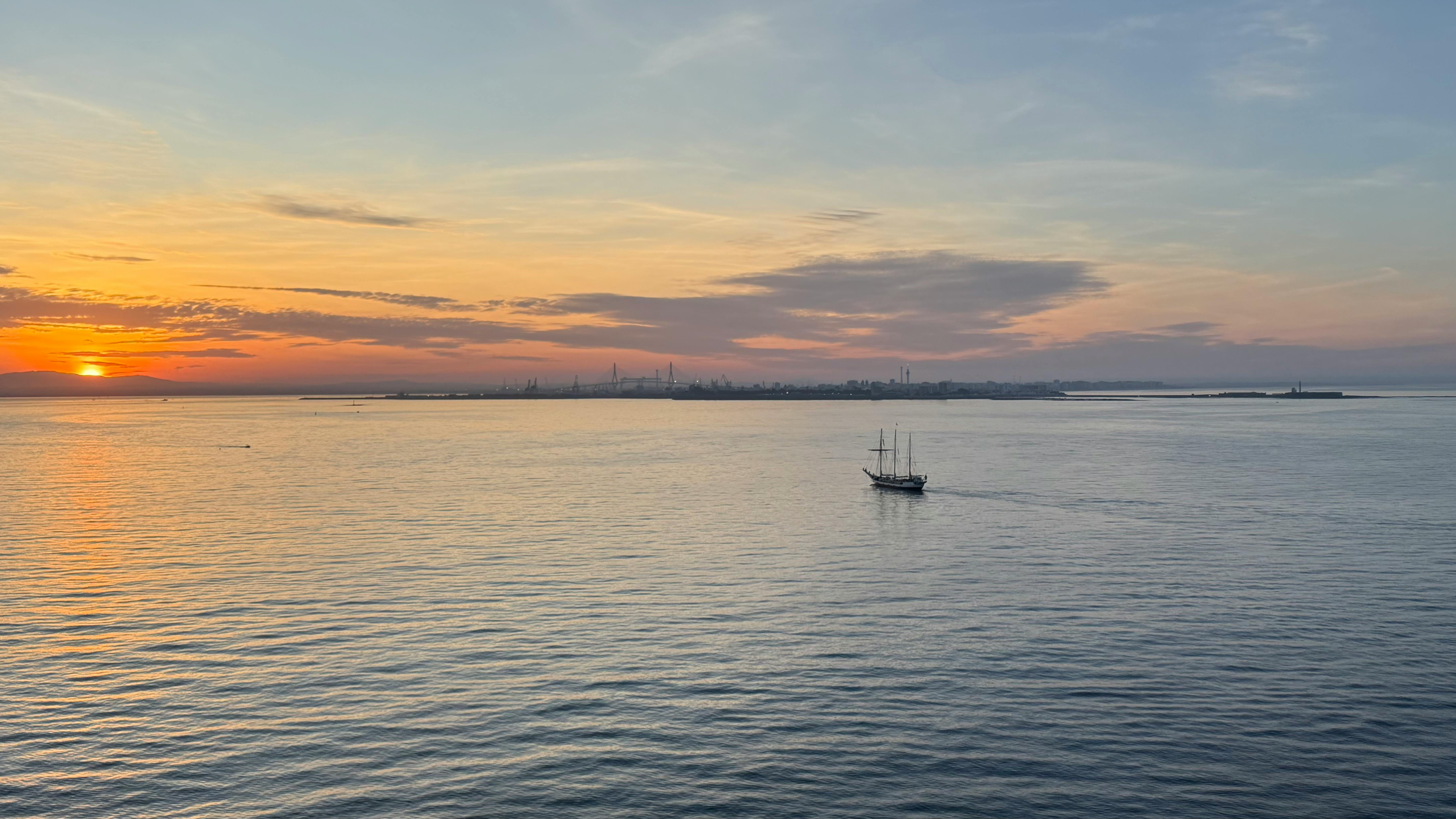 加的斯湾上空，宁静的日落映照着平静的水面，一艘孤独的帆船静静地漂浮着