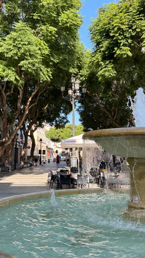Free Serene plaza in Cádiz, Spain, featuring a vibrant fountain and lush trees with seating for an outdoor cafe. Stock Photo