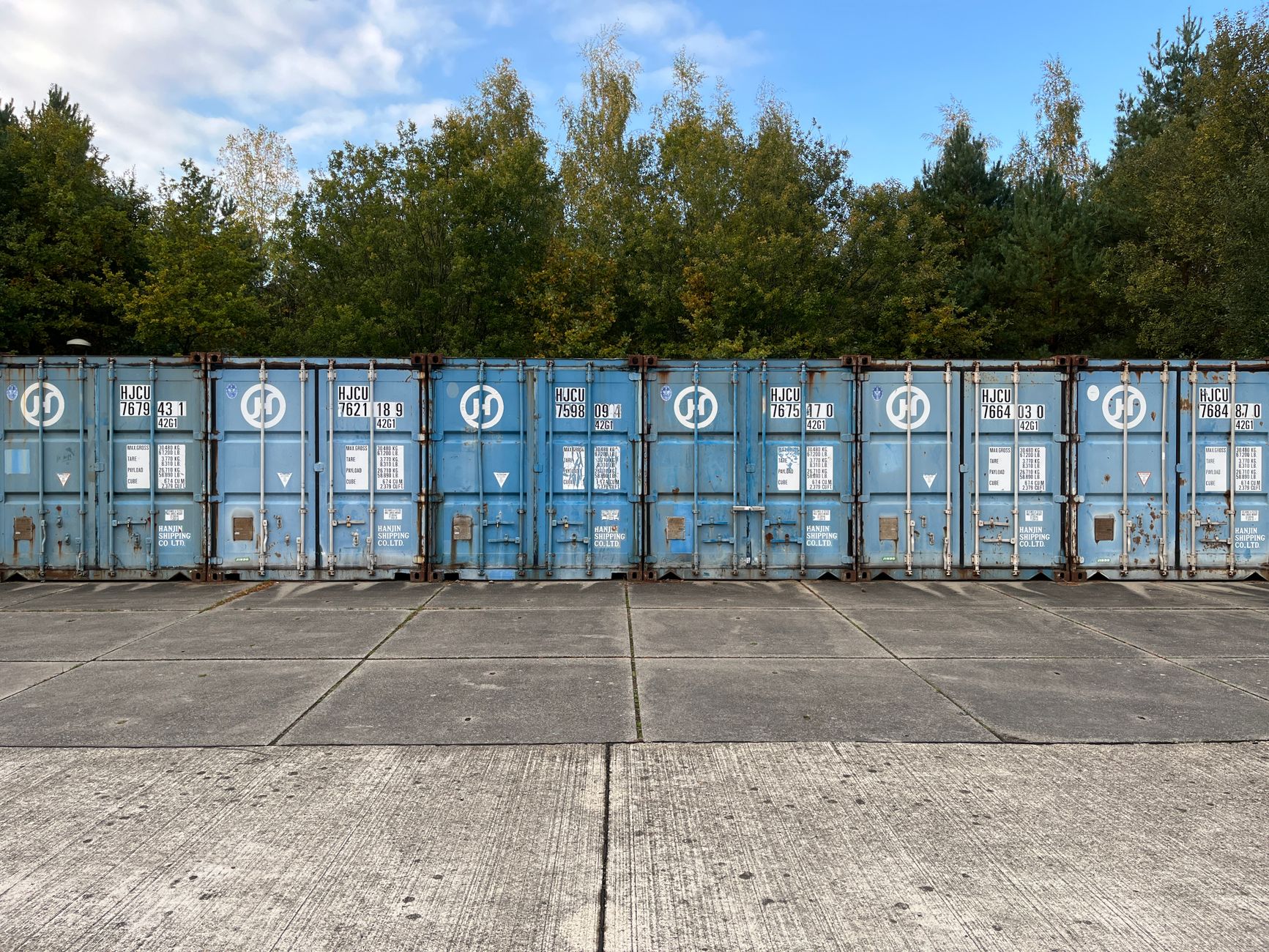 Blue shipping containers lined up outdoors against a forest background.