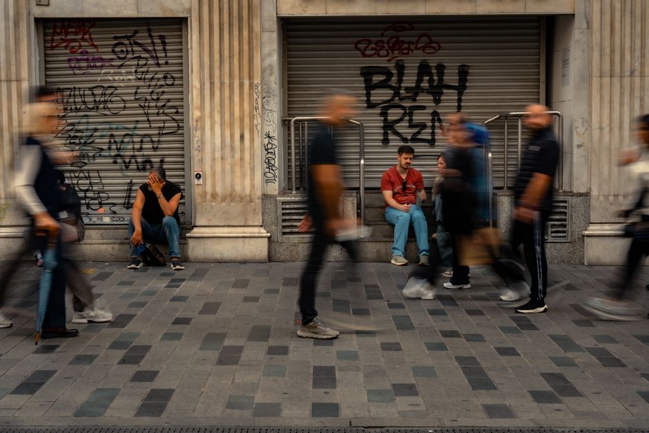 Peatones borrosos pasan junto a un hombre sentado contra una pared graffiteada en un bullicioso entorno urbano.