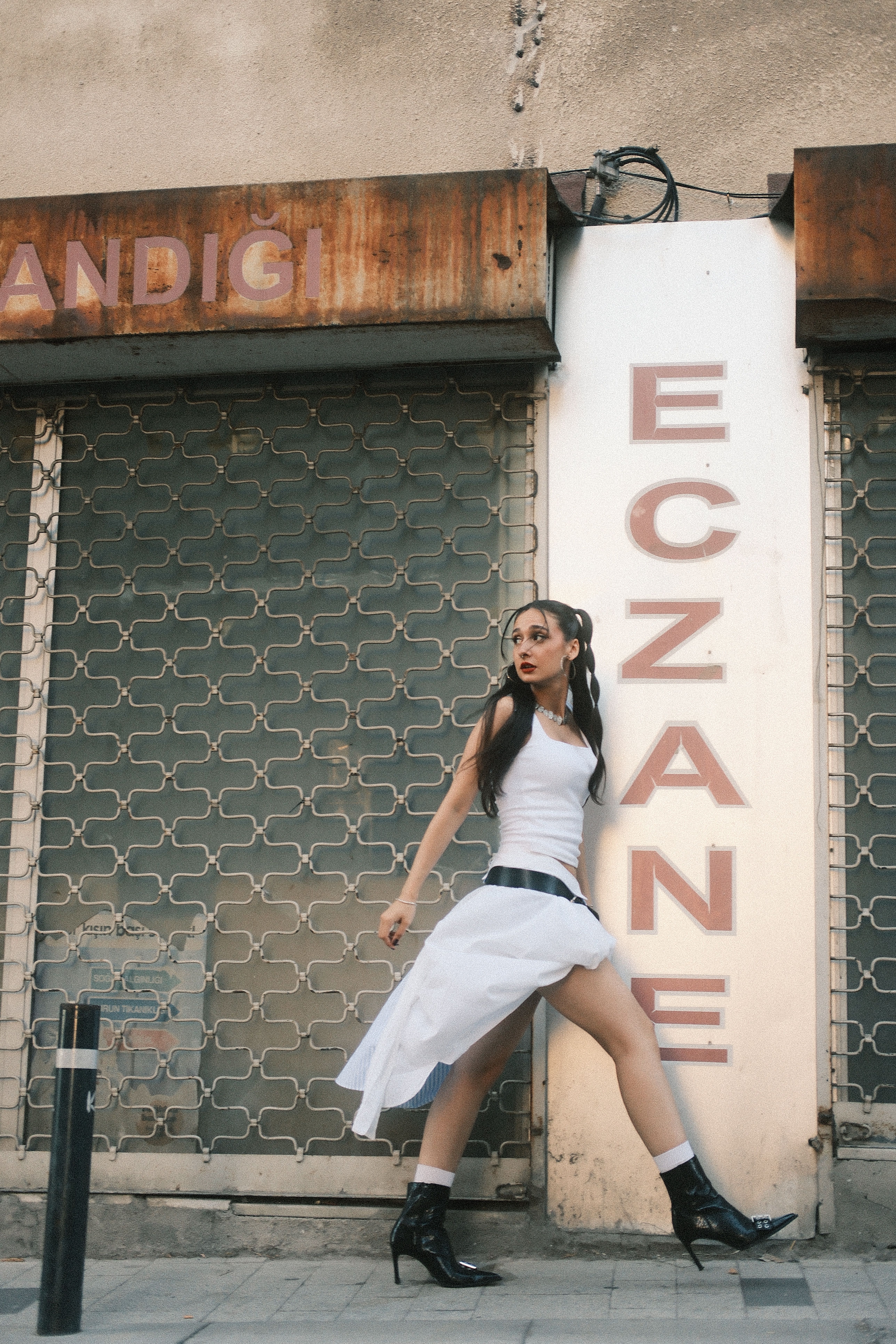Stylish woman in white dress and boots walking by urban background, sign 'Eczane'.