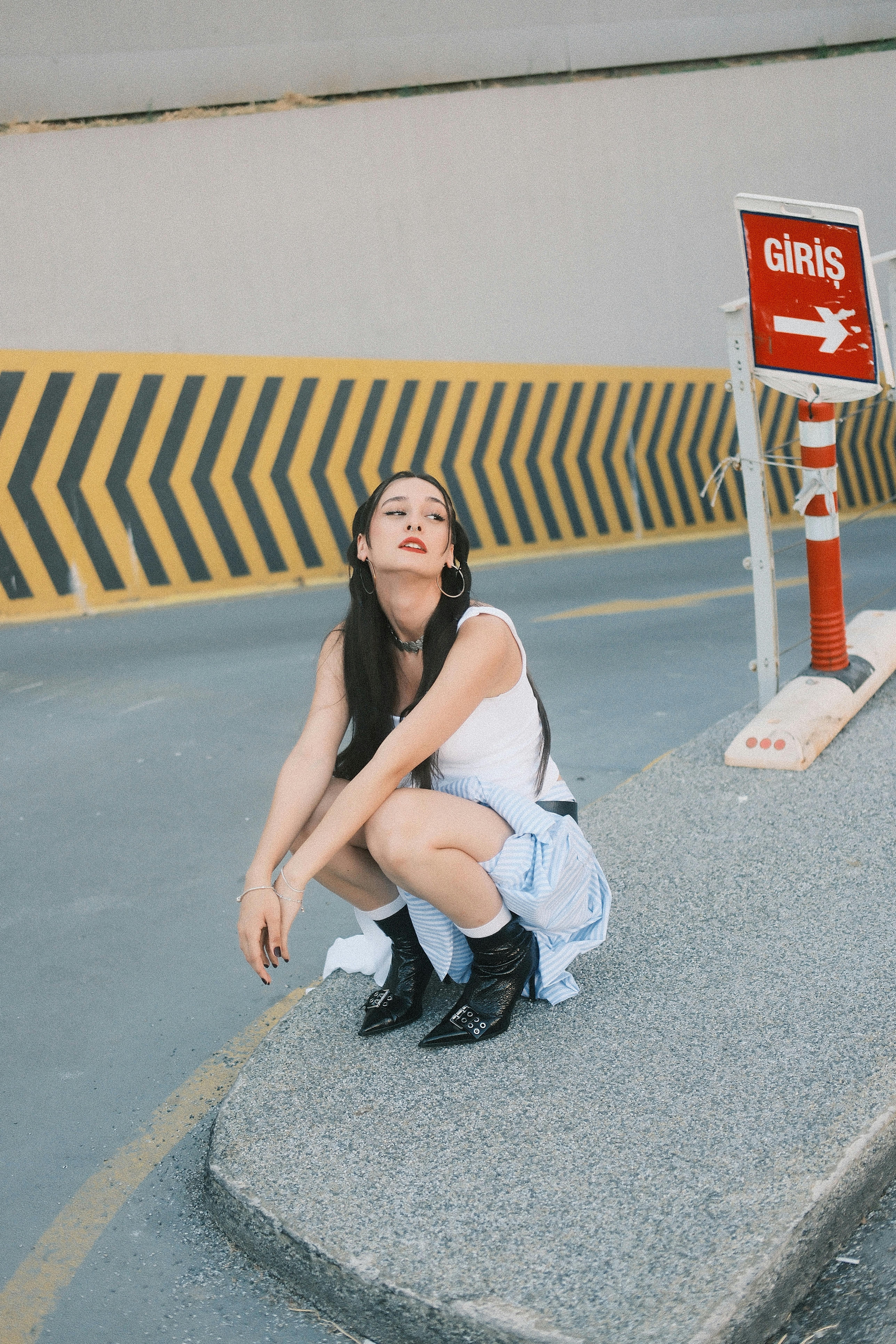 Stylish woman in urban setting, posing beside a 'Giriş' sign on a sunny day.