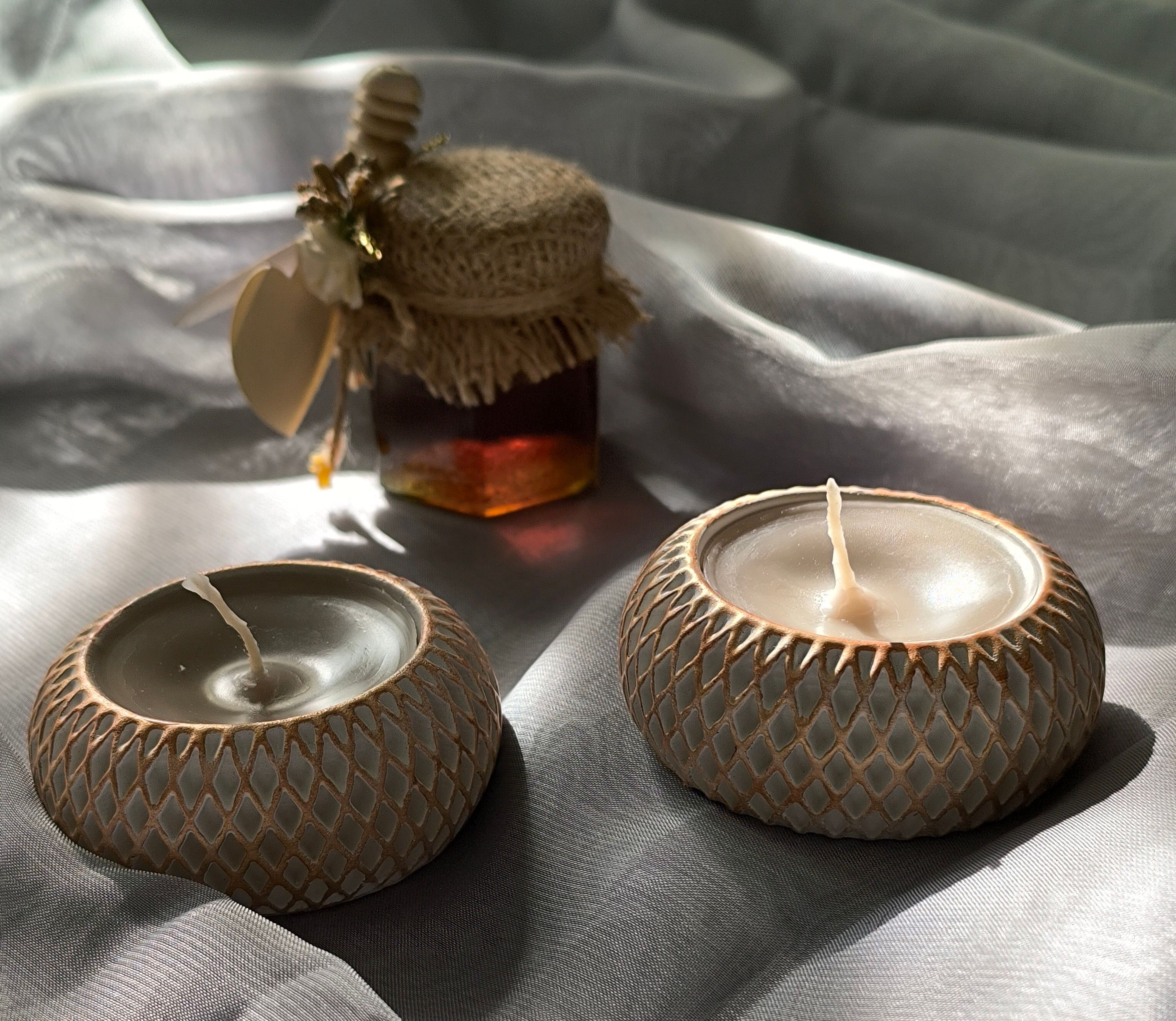 Stylish arrangement of candles and honey jar on silky fabric, lit by soft light.