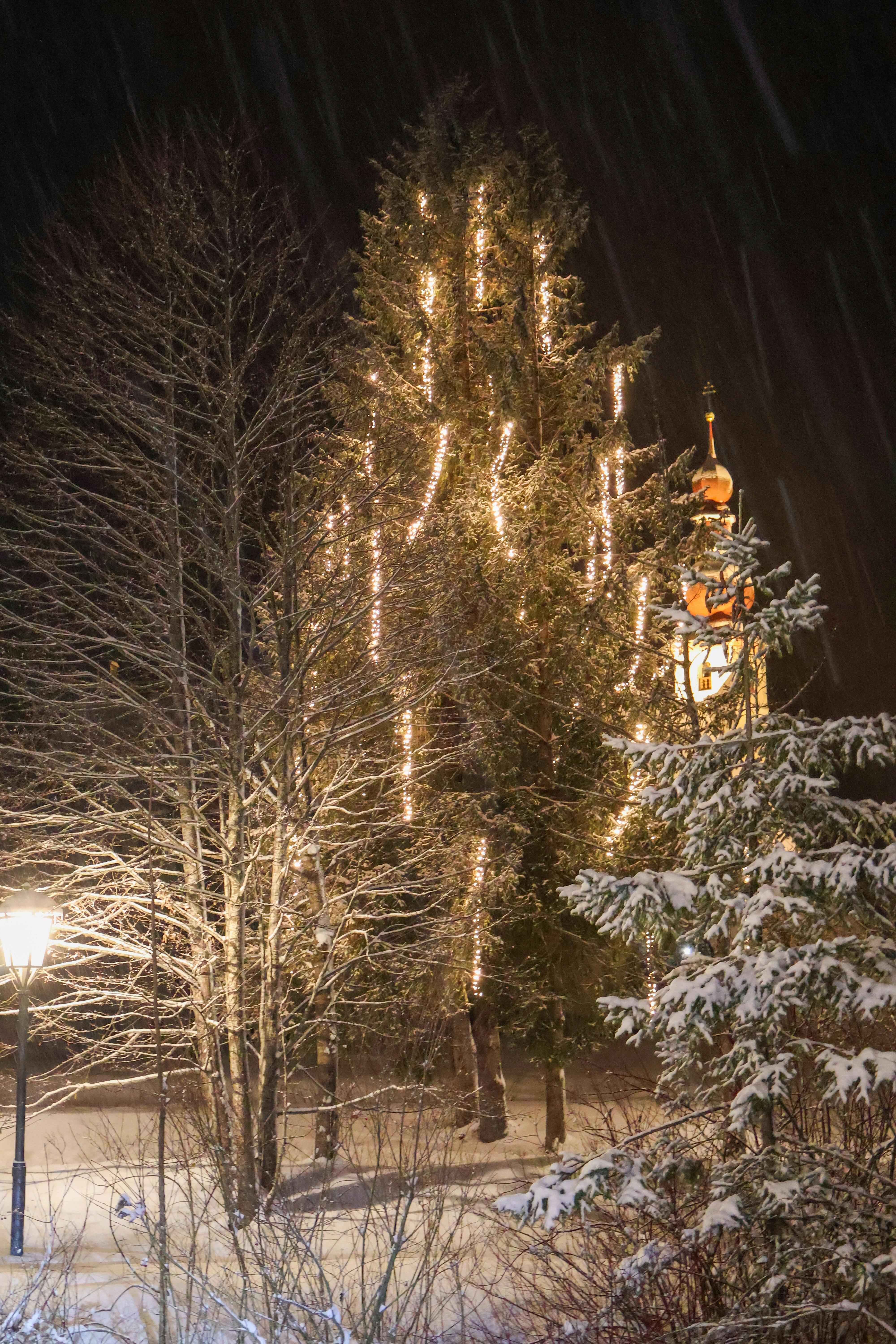 Snowy night scene in Sankt Ulrich am Pillersee, featuring illuminated trees and a church tower.