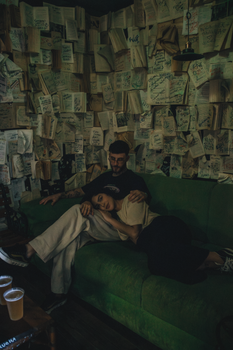 A couple relaxes on a green couch surrounded by open books on walls, creating a cozy atmosphere.