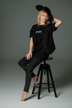 Stylish woman posing barefoot on a stool, wearing a black hat and outfit in a studio setting.