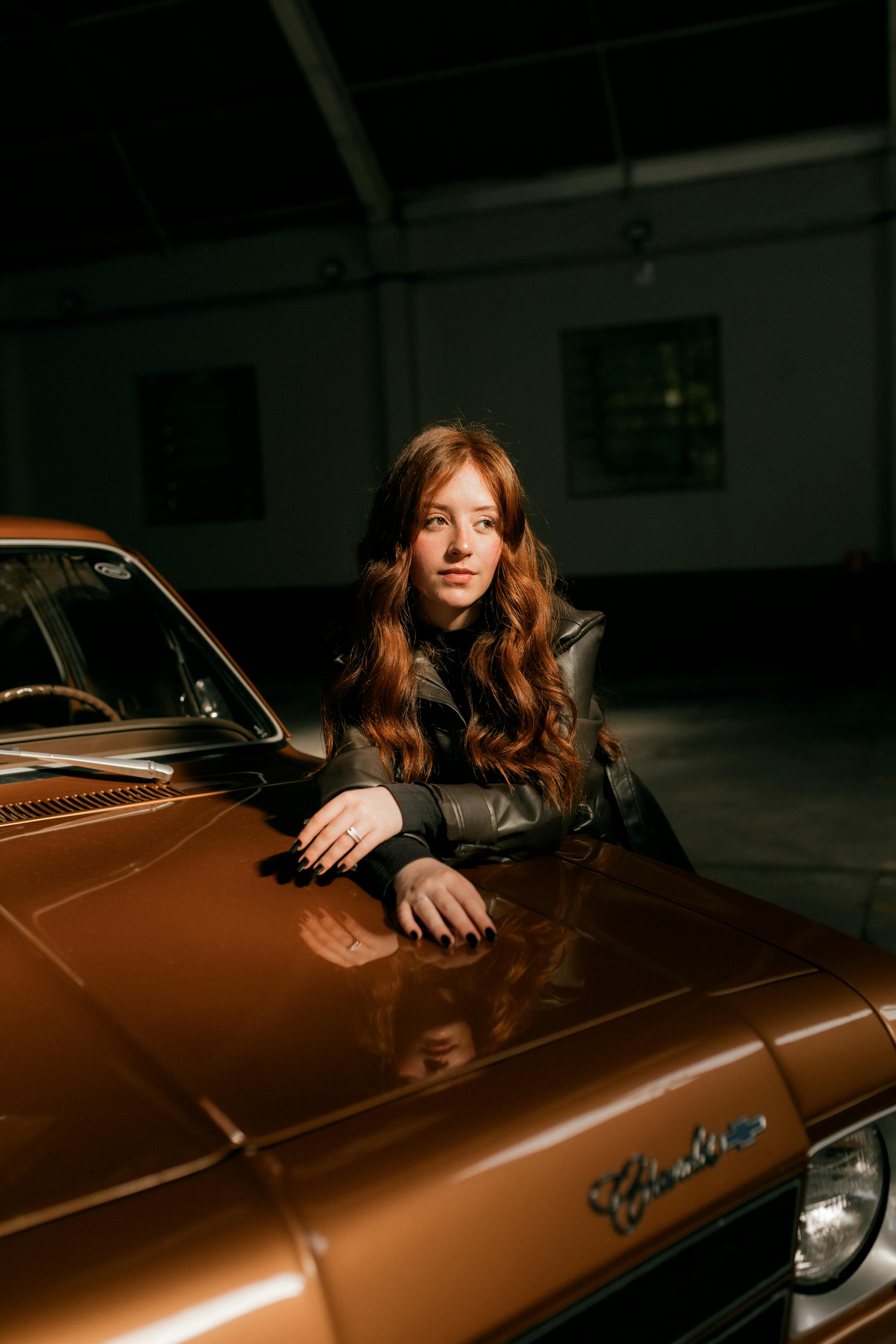 Ücretsiz Loş ışıklı bir garajın içinde eski bir arabaya yaslanmış genç bir kadının portresi. Stok Fotoğraflar