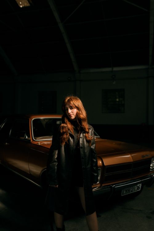 Free Young woman in leather jacket stands by vintage car in dimly lit garage, creating a moody atmosphere. Stock Photo