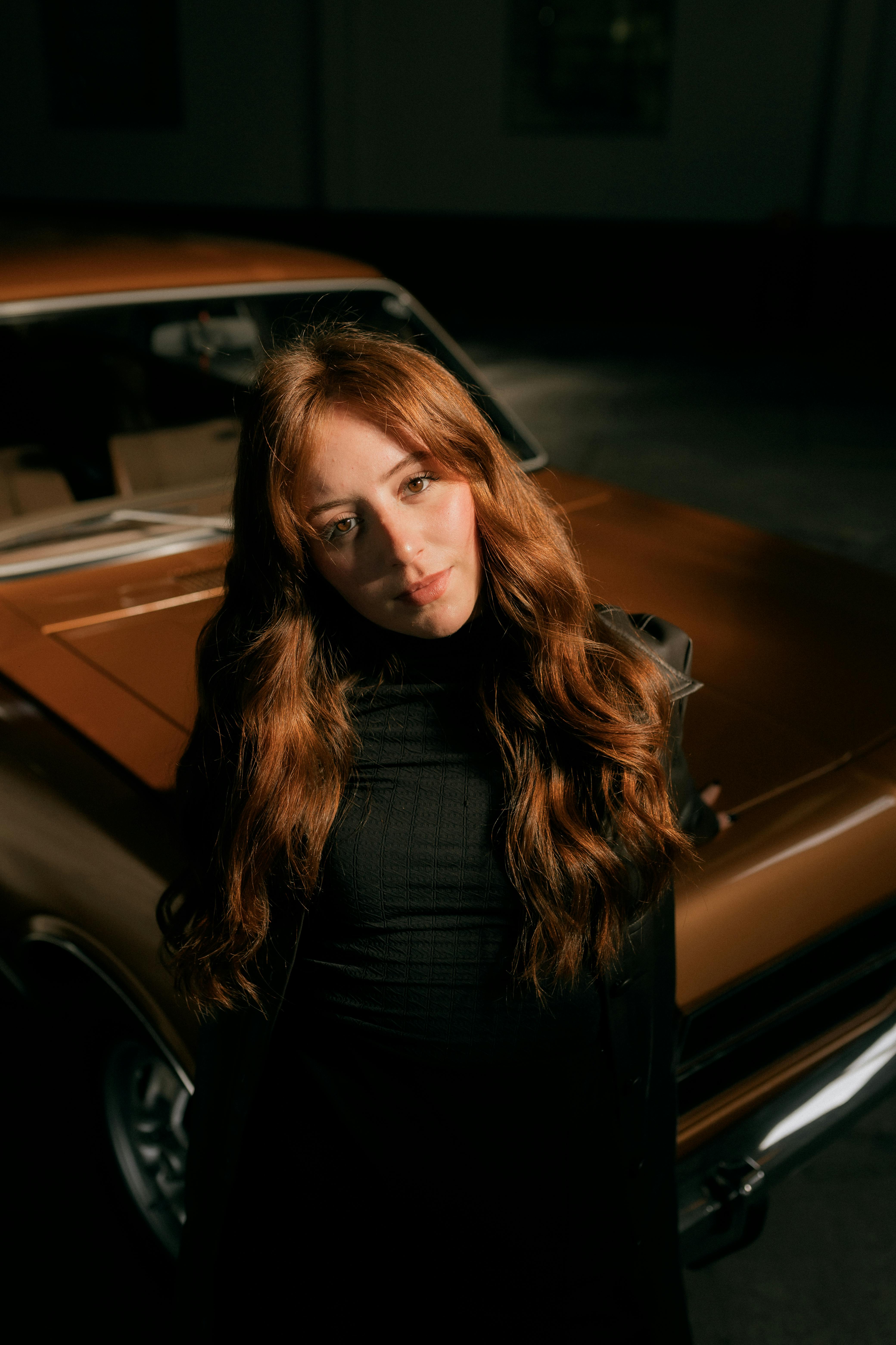 A young woman stands by a classic car in moody lighting, evoking nostalgia.