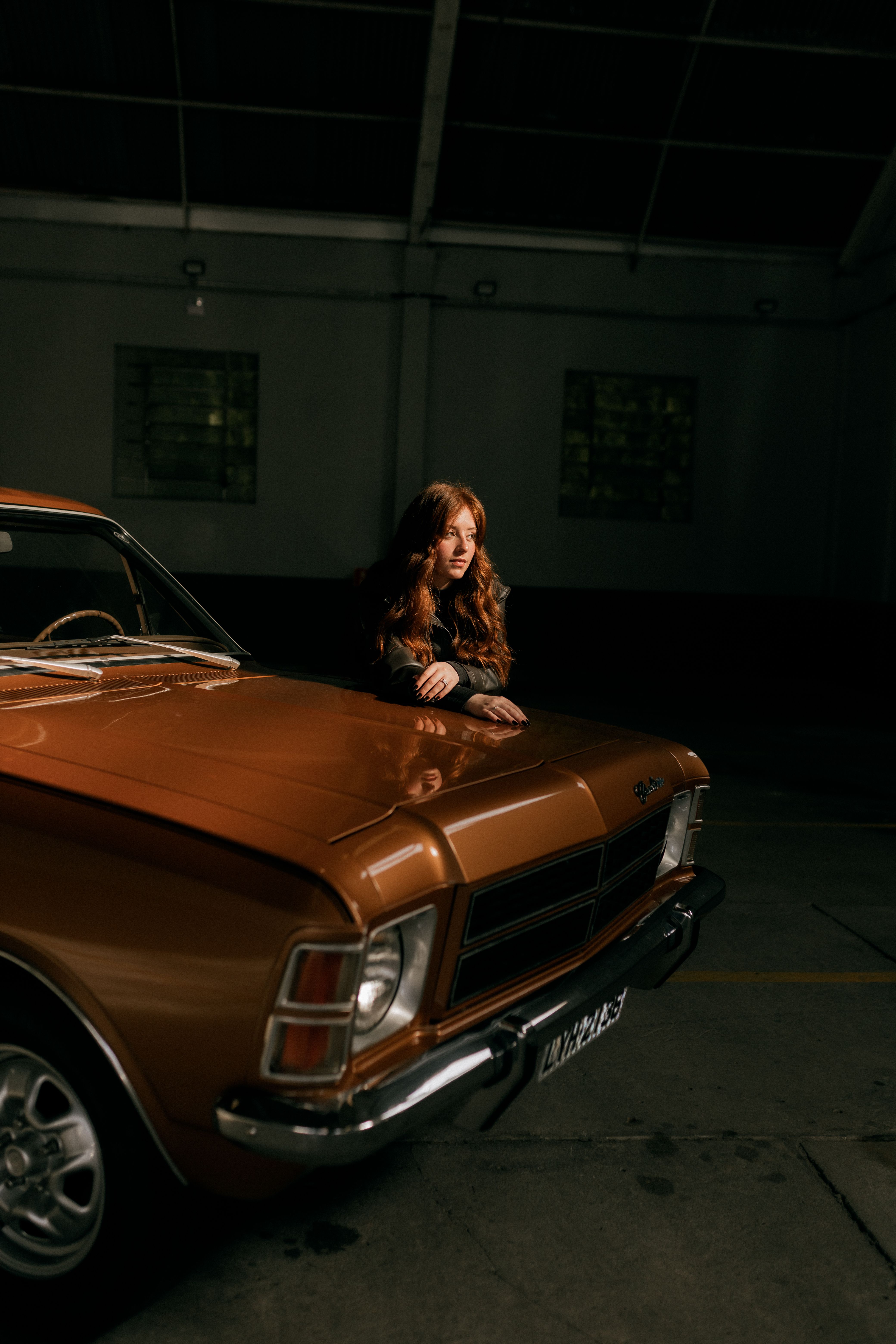 A woman leans on a vintage car indoors, with warm lighting creating a dramatic mood.