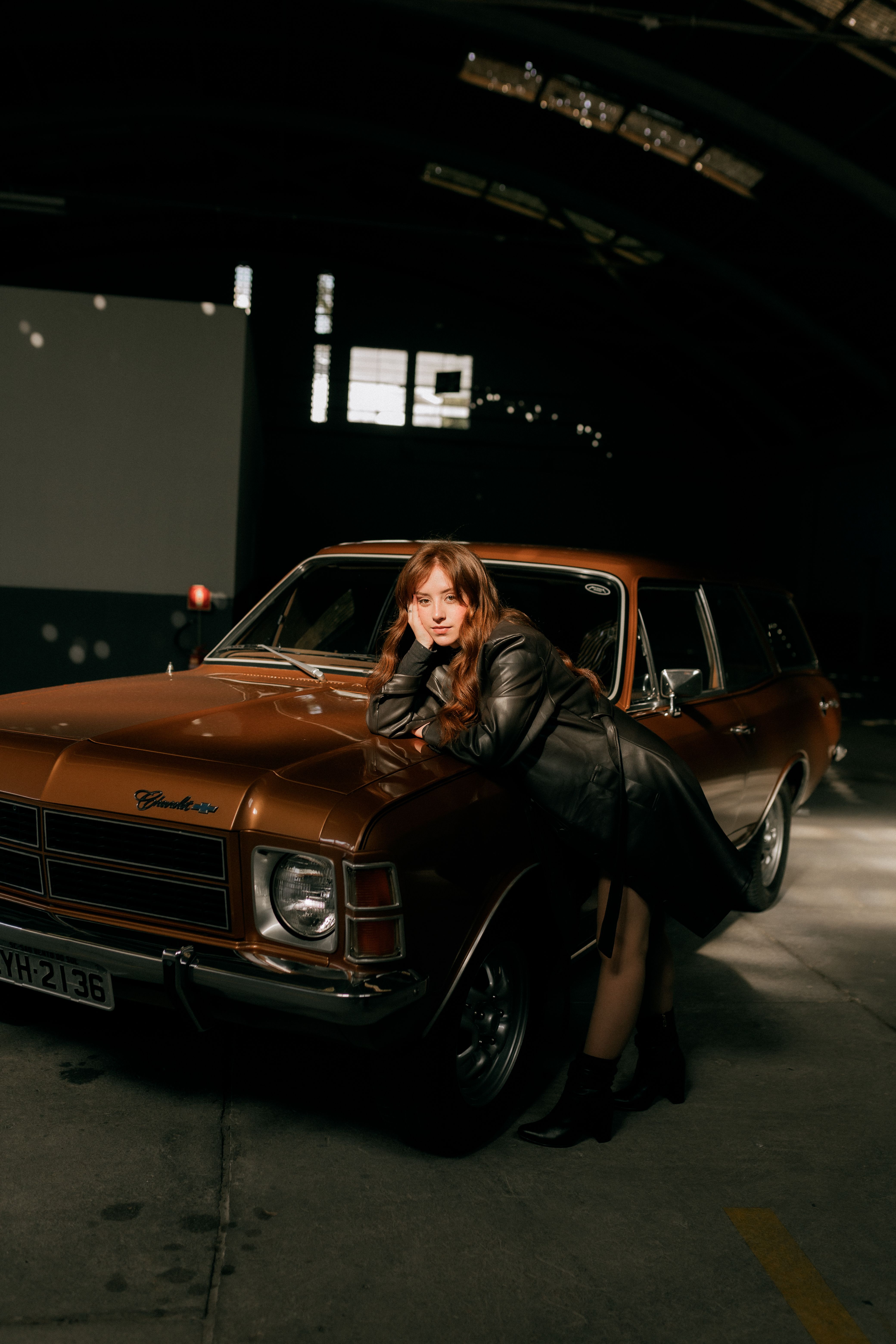 Stylish woman in leather leans on a vintage car in a dimly lit garage.