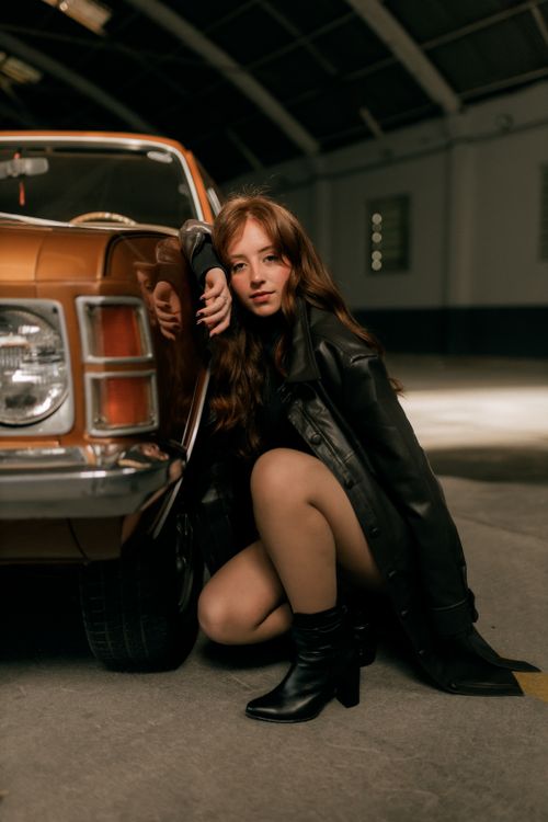 Free Young woman in leather jacket leaning on vintage car in dimly lit garage. Stock Photo