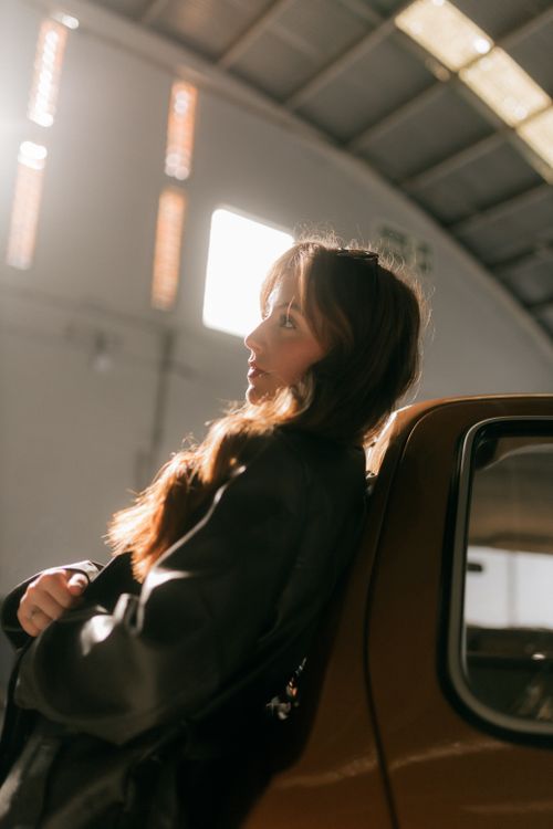 Free A young woman in a leather jacket leans against a car in a sunlit warehouse. Stock Photo