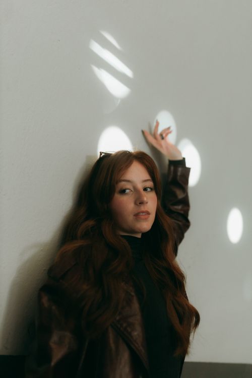 Free Woman in brown jacket in artistic shadows against white wall, serene expression. Stock Photo