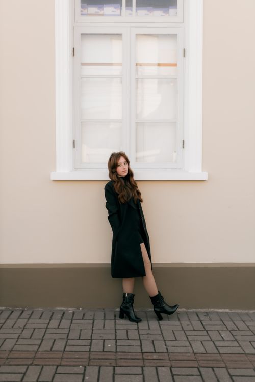 Free Stylish woman in black coat and boots posing by a large window. Elegant fashion portrait. Stock Photo