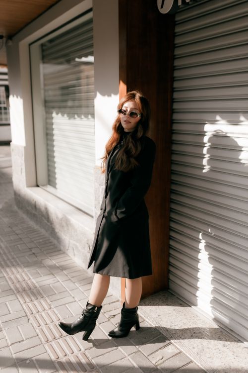 Free Fashionable woman wearing a black coat and sunglasses stands confidently beside a storefront. Stock Photo