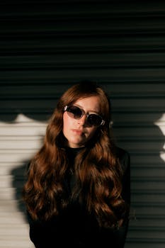 Elegant woman with sunglasses and long hair posing in sunlight. Modern fashion portrait.
