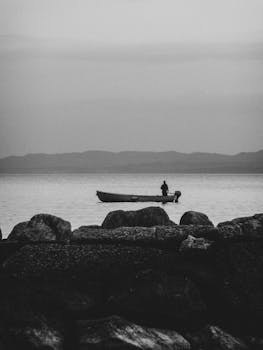 A lone fisherman in a boat on a calm lake with distant mountains.
