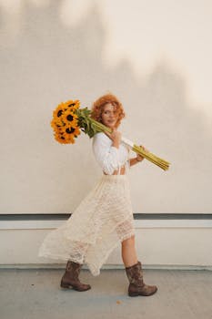 Charming woman with curly hair in a lace dress carries sunflowers, embodying nature's beauty.