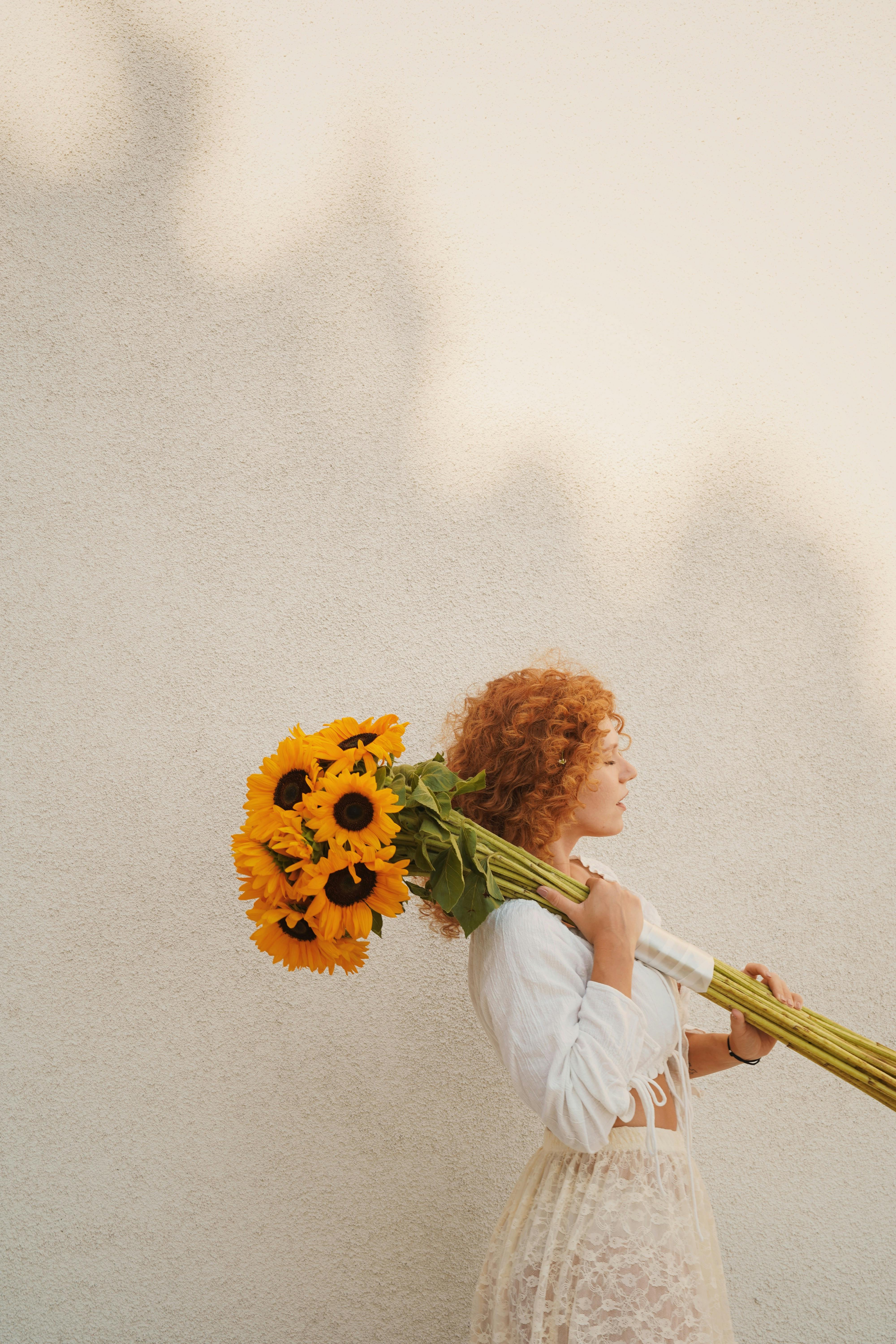 Side view of a woman with curly hair carrying a bouquet of sunflowers against a textured wall, embodying a rustic vibe.