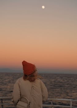 Woman in a beanie observing a serene sunset over the ocean from a boat.