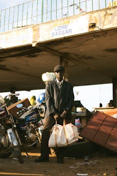 Man in suit holding bags with motorbikes at outdoor market under bridge.