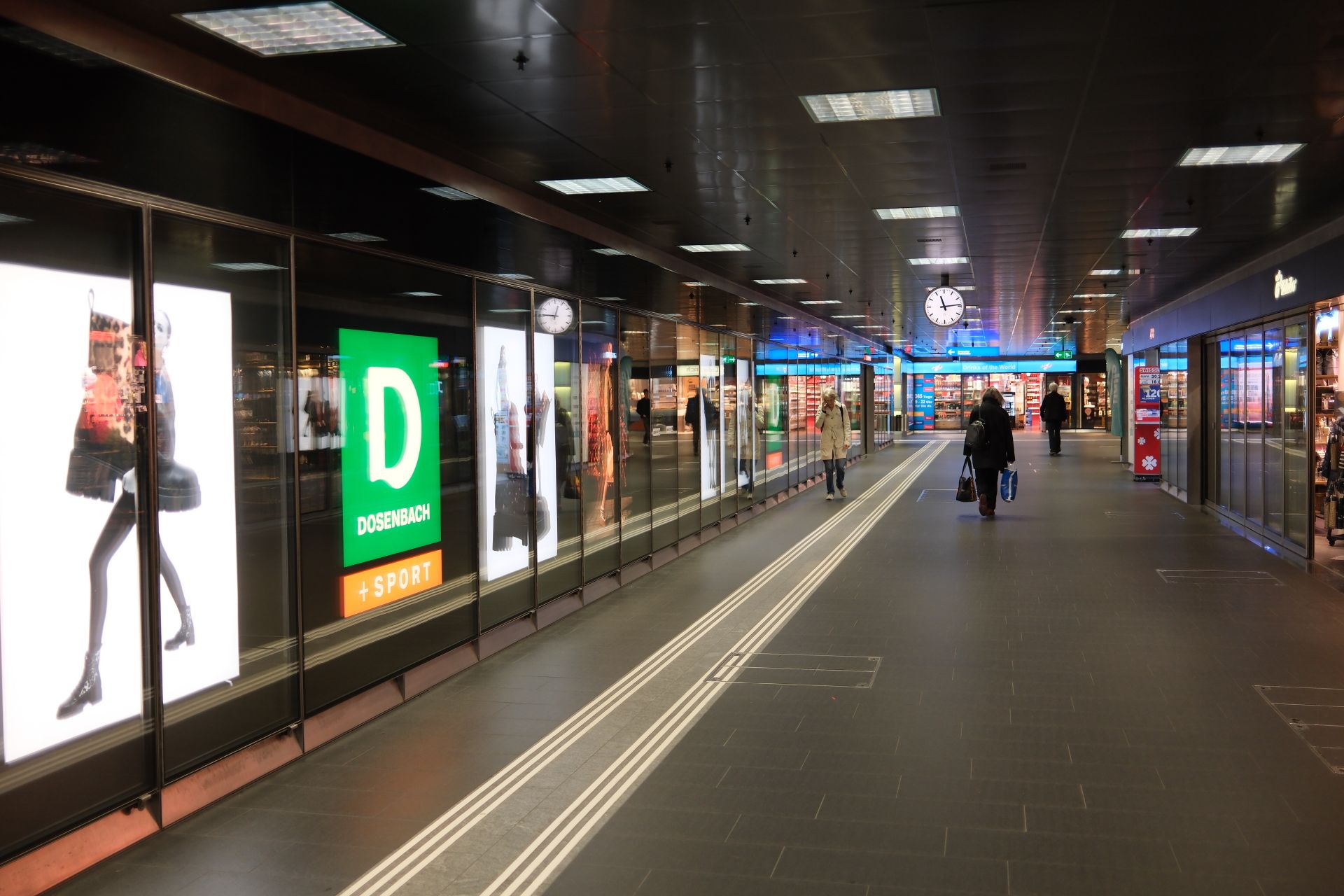 Wide view of a busy shopping mall corridor with people, shops, and a prominent clock.