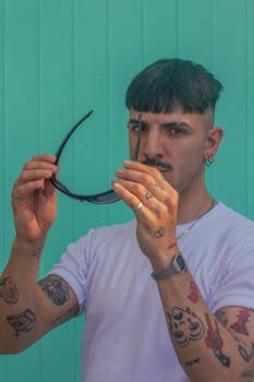 Stylish tattooed man holding sunglasses with a teal wall backdrop, exuding urban vibes.