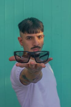A young man presenting sunglasses with a teal wall background in Argentina.