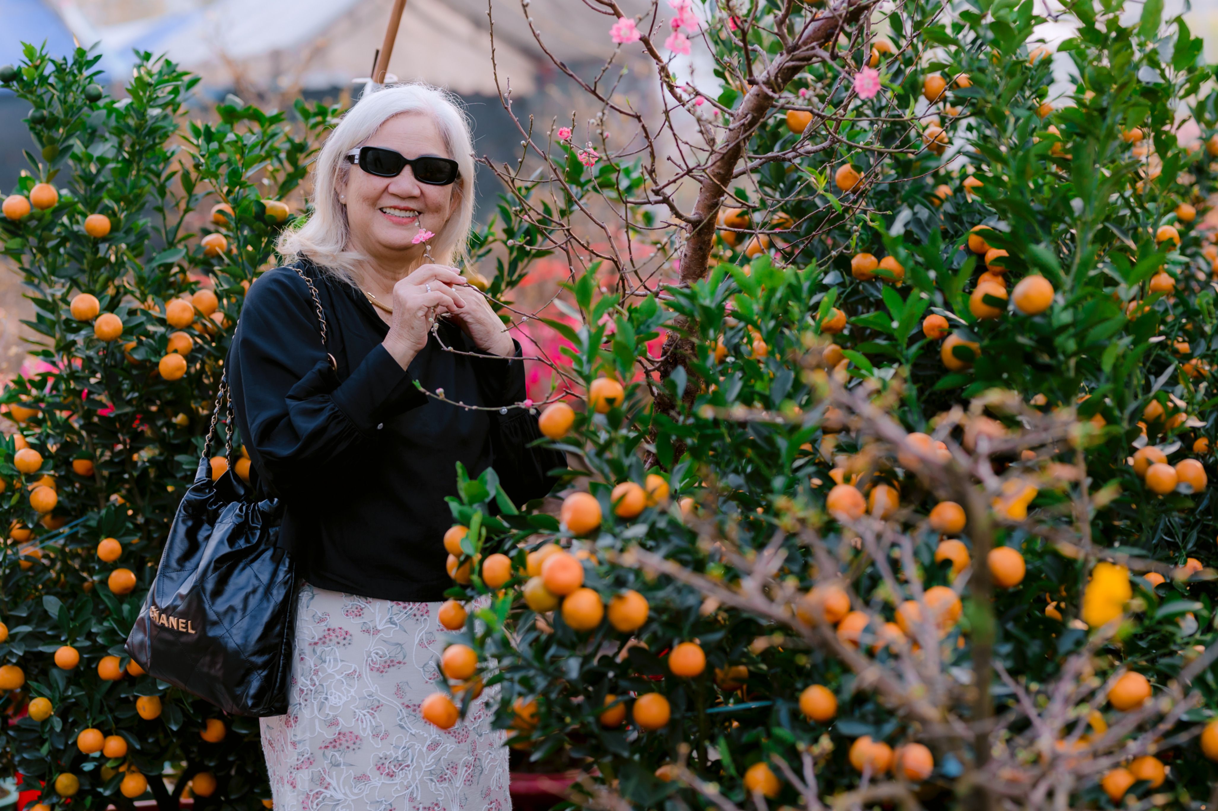 Senior woman with sunglasses smiling in a vibrant outdoor garden surrounded by orange trees.