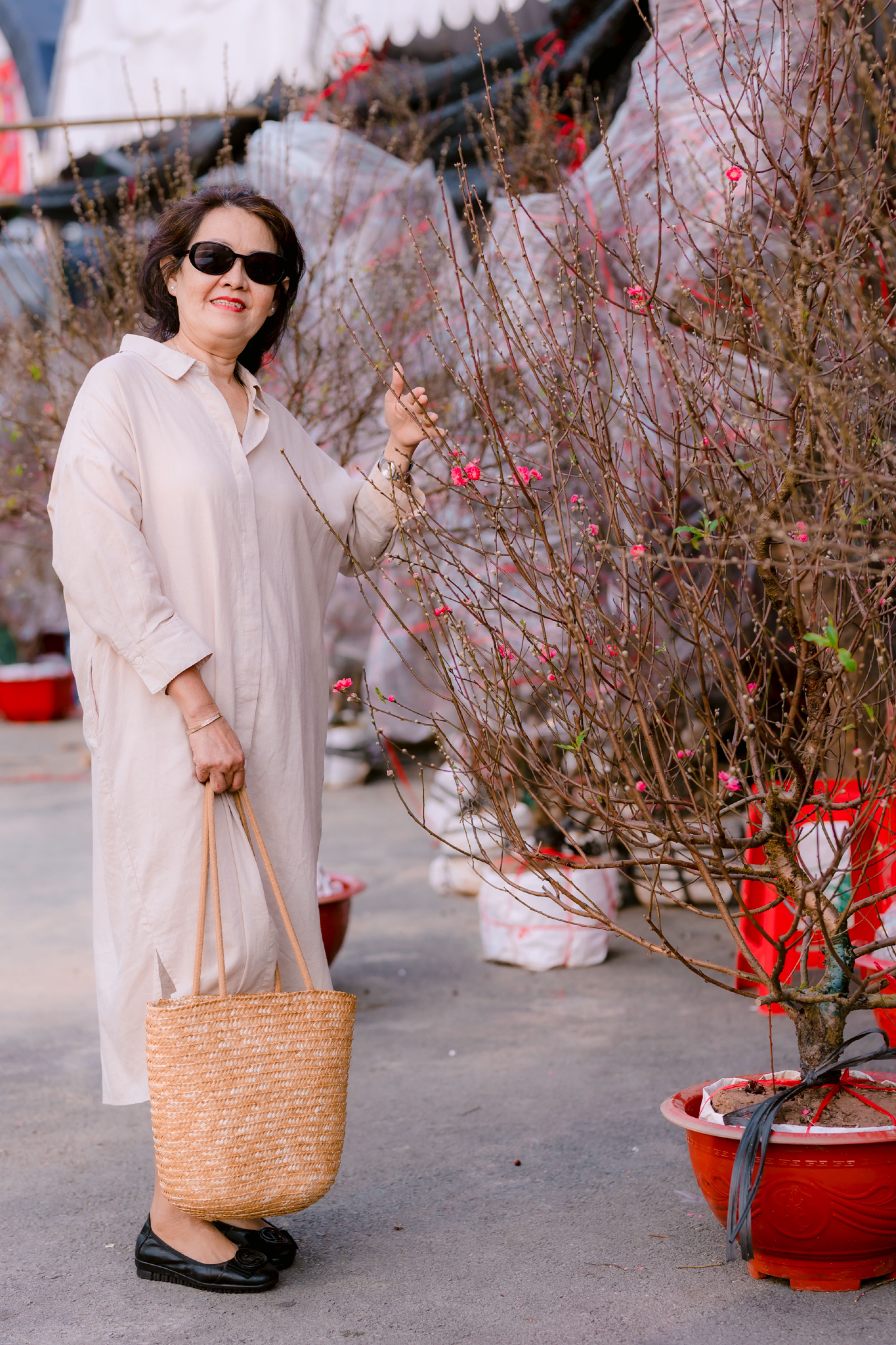 A woman in sunglasses and a long dress admires blooming trees in pots outdoors, carrying a woven bag.