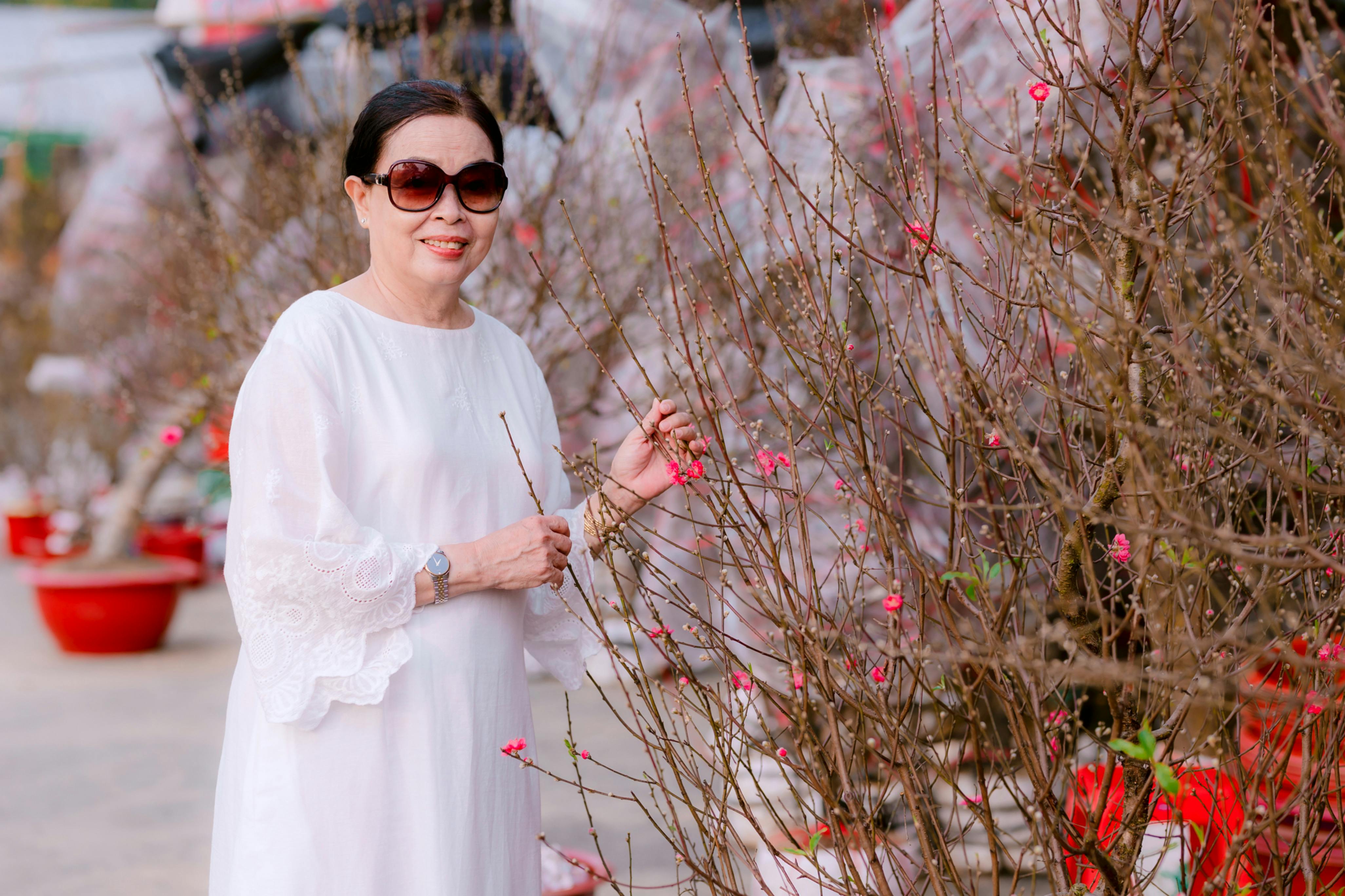 Senior woman smiling and enjoying blooming peach trees outdoors, wearing sunglasses.