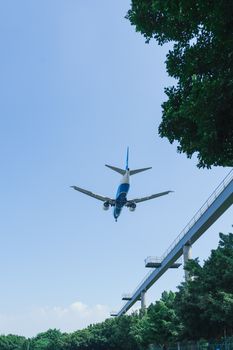 A low-flying airplane captured from below against a clear blue sky, near green trees and a structure.