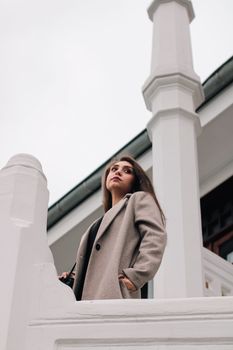 Woman in a beige coat standing by white architecture, looking thoughtfully upwards.