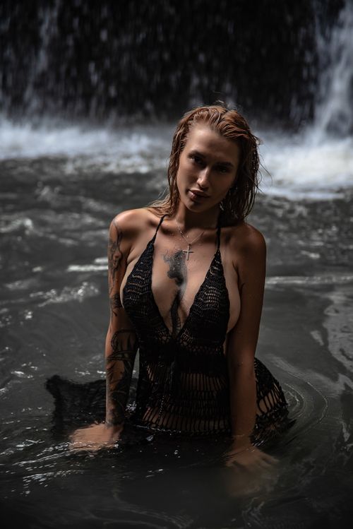 Free A woman with tattoos poses confidently in a dark pool beneath a waterfall, exuding strength and elegance. Stock Photo