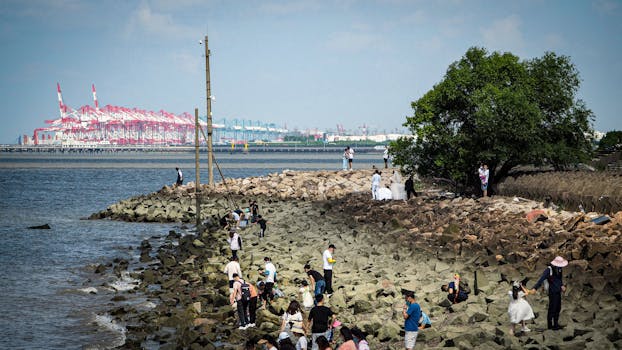 人们在晴朗的天空下探索繁忙的集装箱港口附近的岩石海岸