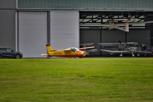 Free A vibrant yellow and orange small plane parked outside a large hangar on a grassy field. Stock Photo