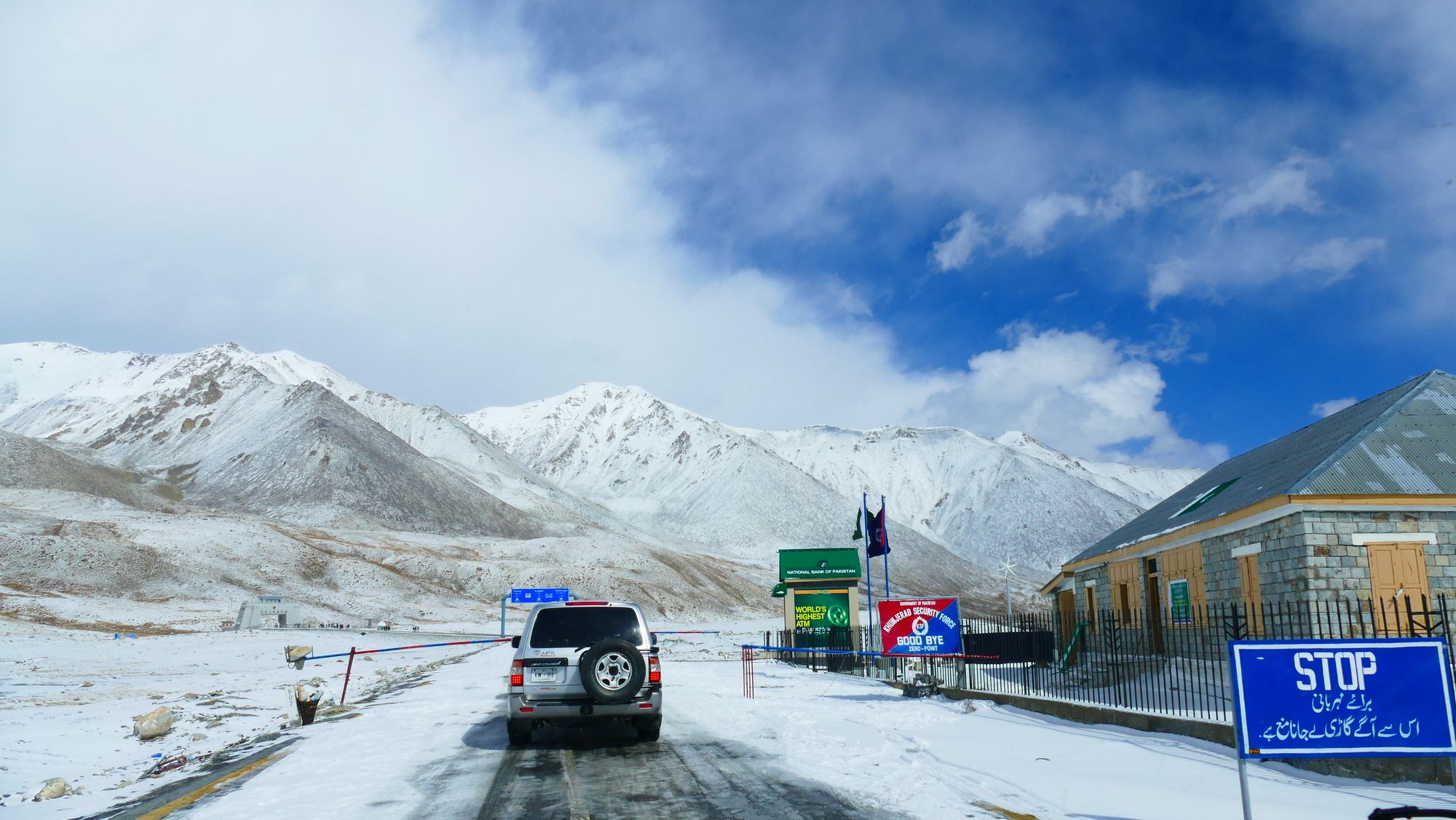 A vehicle approaches a snow-covered mountain pass with border signs and snowy peaks in the background.
