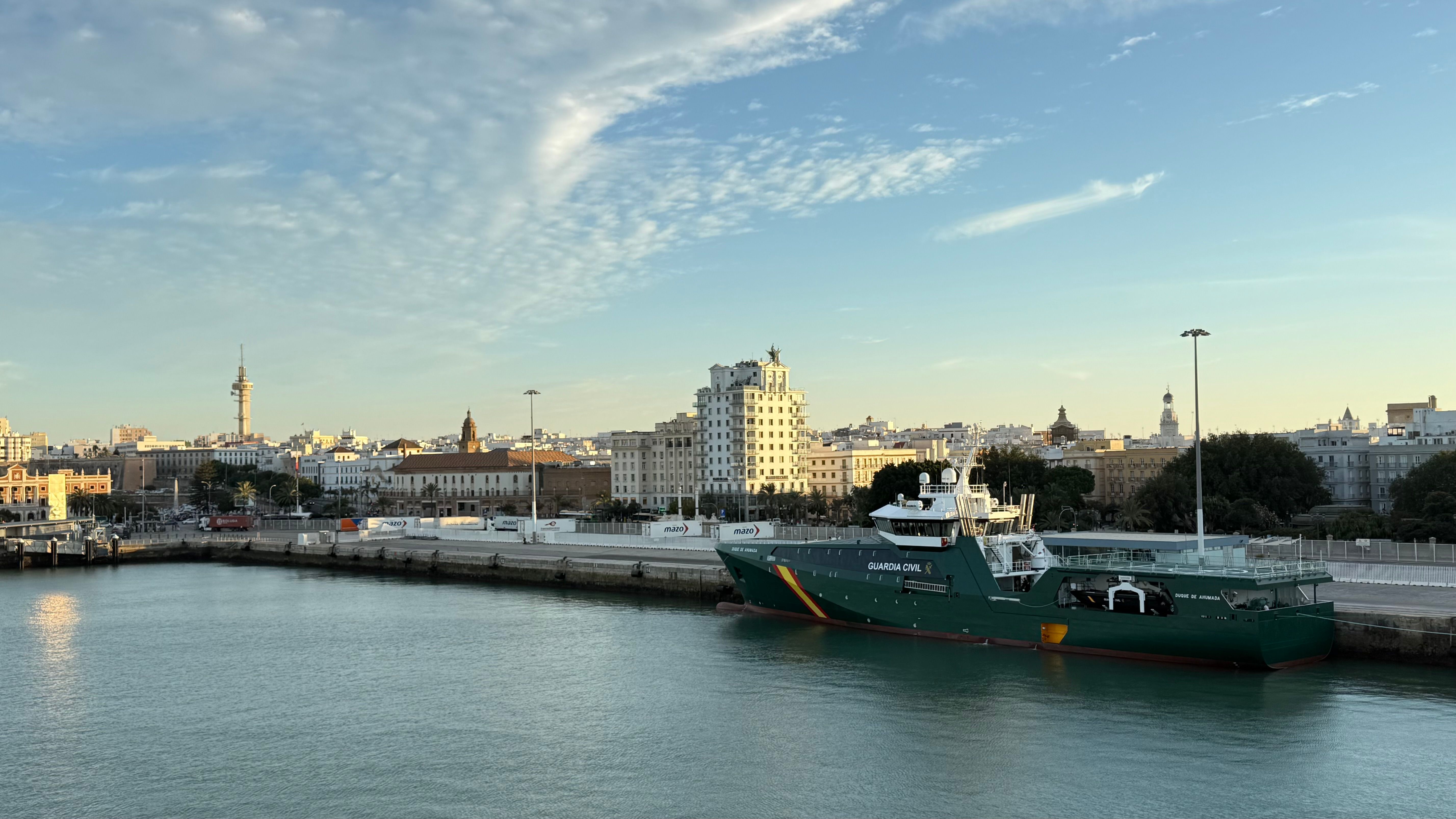 Guardia Civil patrol vessel moored at a Spanish port with the city skyline and calm waterfront in the background