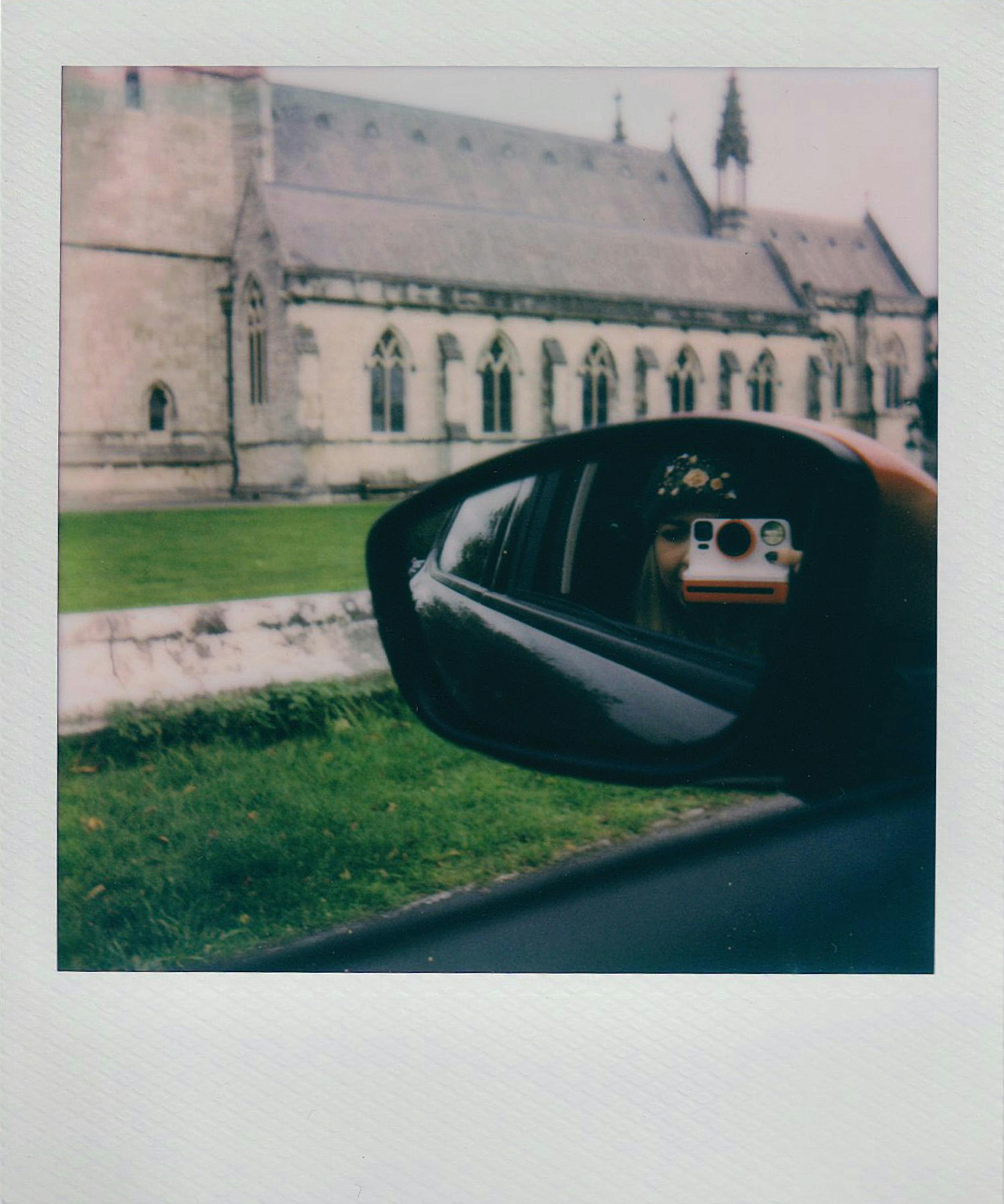 A woman holding a polaroid camera is reflected in a car mirror with a church in the background.