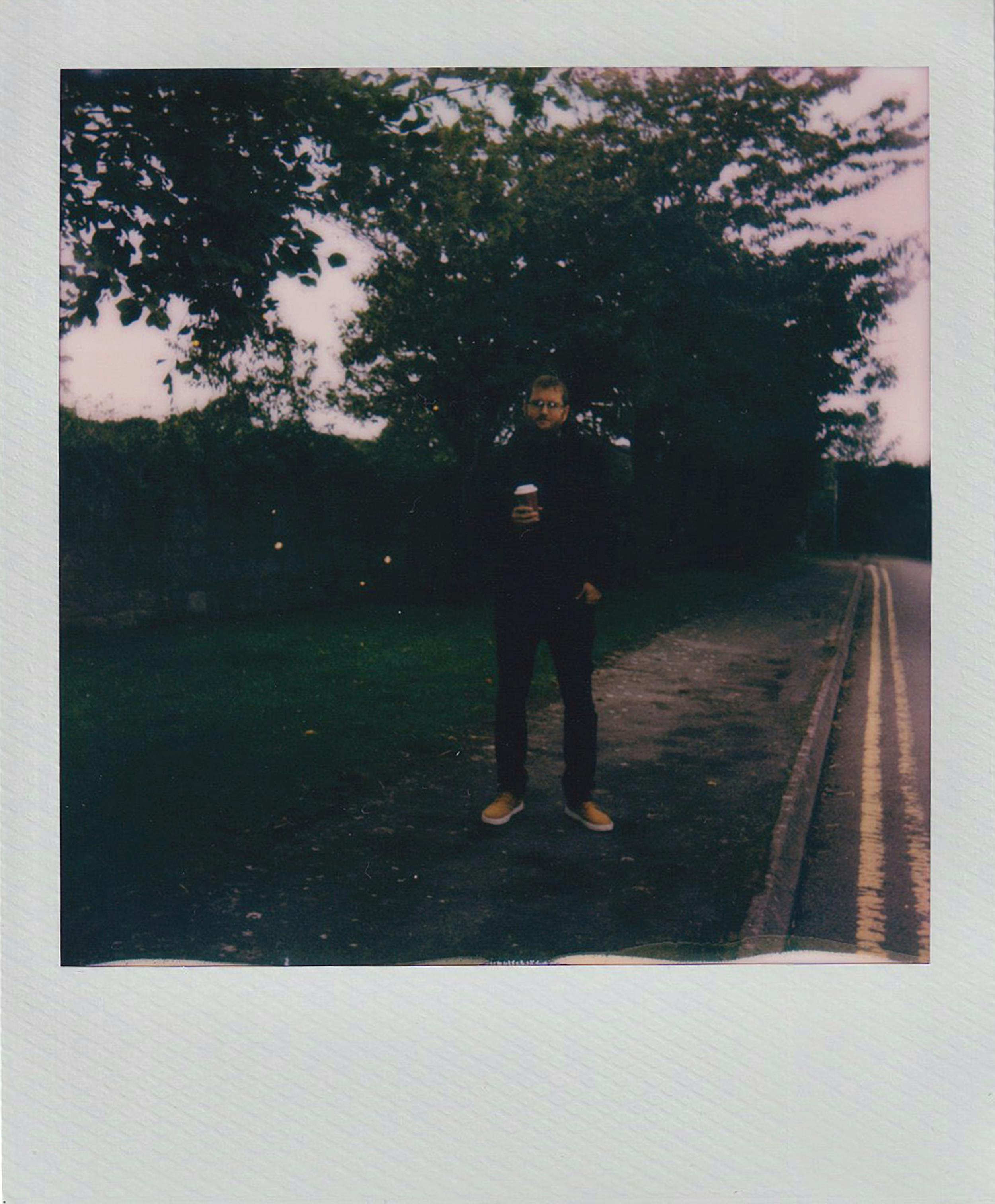 A person standing on a path beside trees with a vintage Polaroid effect.