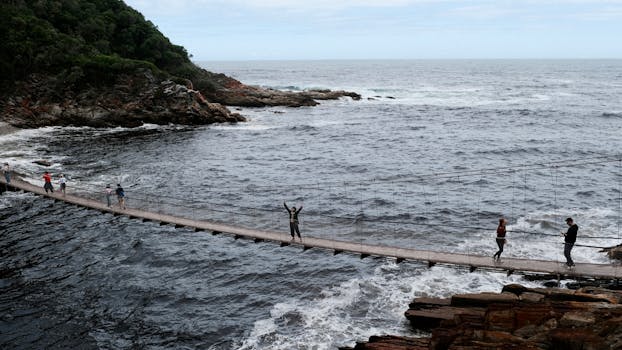A scenic suspension bridge with people crossing above turbulent ocean waves.